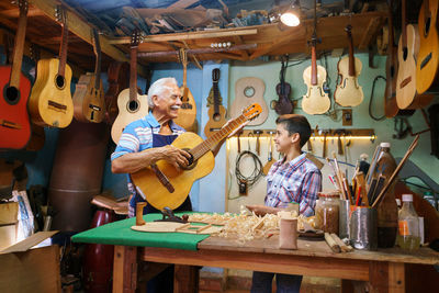 Senior man playing guitar while standing with boy in workshop