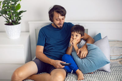 Bearded father and son are sitting on the bed in t-shirts and watching a football match on tv