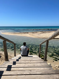 Rear view of boy walking on pier at beach against clear blue sky