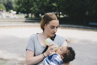 Young mother feeding baby boy from bottle at park