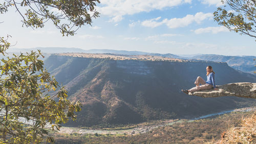 Woman resting on cliff while looking at mountains against sky