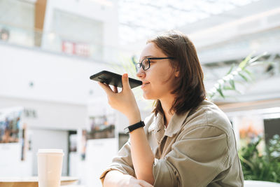 Young brunette teenager girl student in glasses using mobile phone sent voice message at cafe