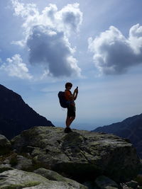 Side view of silhouette boy using smart phone while standing on rock against sky