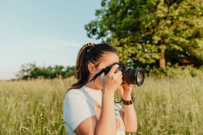 Young woman standing on meadow, using camera and taking photos at golden hour