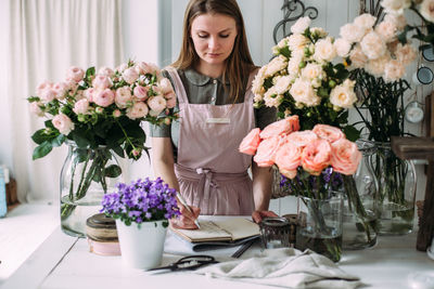 A young girl florist writes in a craft notebook in a store among the flowers.