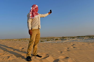 Man with covered face taking selfie while standing on sand at beach