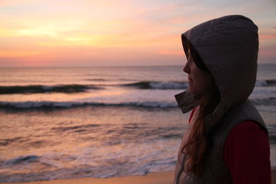 Side view of hooded woman standing at sea shore during sunset