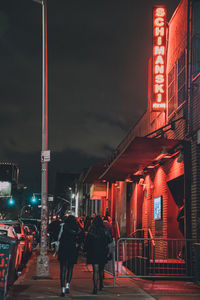 Rear view of people walking on illuminated street amidst buildings at night