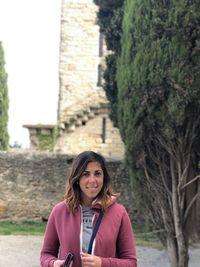 Portrait of smiling young woman standing against trees and building