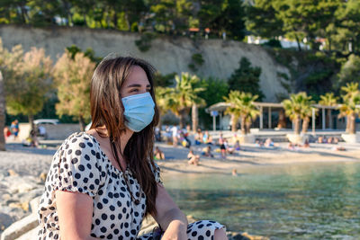 Young woman wearing surgical mask, sitting and relaxing on beach in summer.