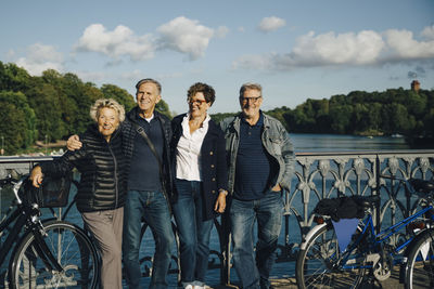 Happy senior friends standing at railing of bridge against sky