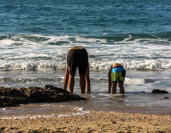 Father and son bending at sea shore against clear sky