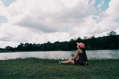 Side view of woman sitting on grass by lake against sky