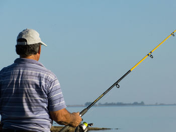 Rear view of man fishing in sea against clear blue sky