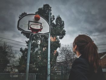 Low angle view of woman looking at basketball hoop against cloudy sky