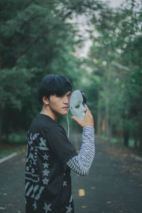 Portrait of young man holding mask while standing against trees