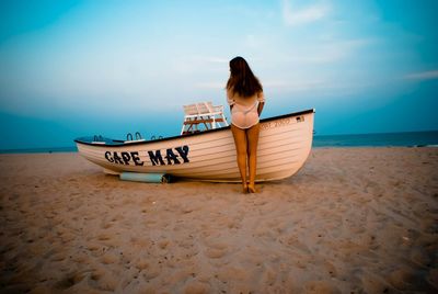 Rear view of sensuous woman standing by boat at beach against sky