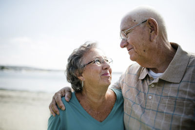 Happy senior couple looking each other face to face while standing at beach against sky