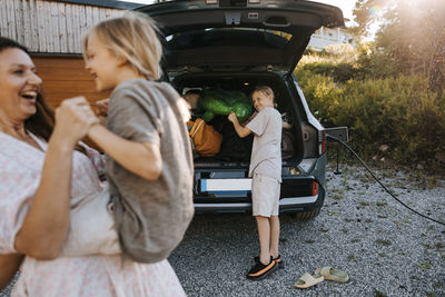 Boy loading car trunk and looking at mother playing with sibling on driveway