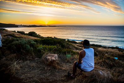 Man sitting at beach against sky during sunset