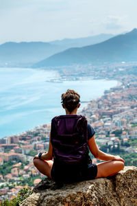 Side view of woman sitting on cliff by sea against sky