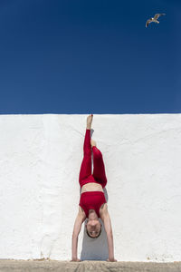 Woman doing a handstand against a white wall with a blue sky wearing red.