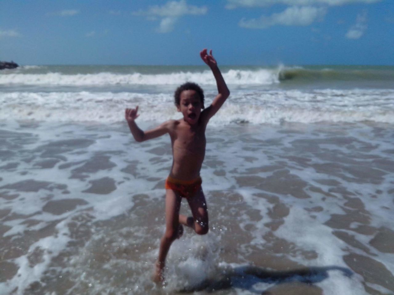 shirtless, one person, beach, real people, sea, arms raised, leisure activity, looking at camera, sky, full length, happiness, water, smiling, fun, portrait, outdoors, day, enjoyment, motion, nature, young adult, front view, vacations, standing, cheerful, lifestyles, young men, childhood, sand, horizon over water, boys, people