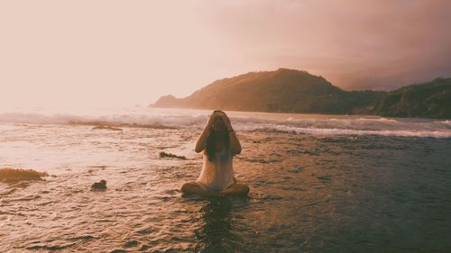 Woman sitting in sea against sky during sunset