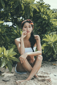 Portrait of woman in bikini sitting on sand by plants at beach