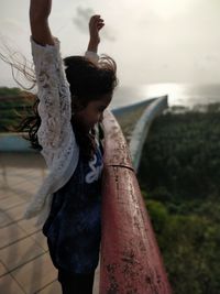 Side view of girl with arms raised standing at railing against sky during sunset
