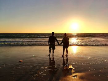 Rear view of silhouette couple holding hands while standing at beach against clear sky