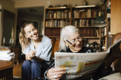 Smiling senior woman reading newspaper while happy female caregiver in background at home