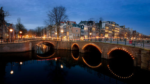 Illuminated bridge over river by buildings against sky at night