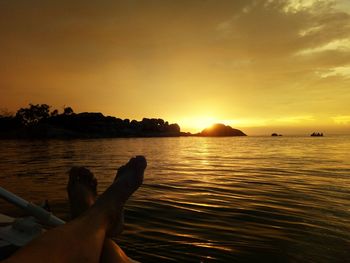 Low section of person relaxing by lake against sky during sunset