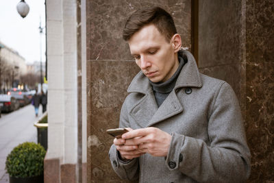 Portrait caucasian young man using his mobile phone in gray coat stands