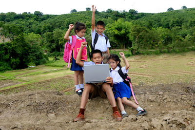 Cheerful siblings looking at brother using laptop while sitting on field against trees