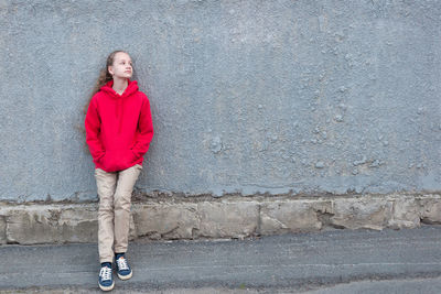 A young blonde stands in a red hoodie, blue sneakers and beige trousers against a gray concrete wall