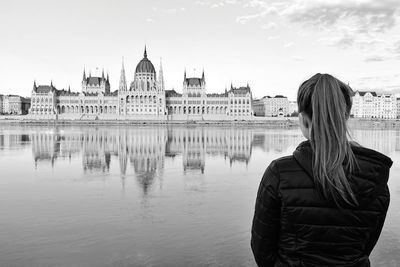 Rear view of woman standing by river against sky in city