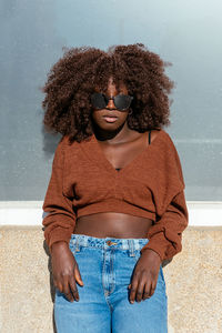 Confident black female with curly hair wearing trendy top with denim and sunglasses standing against wall and looking at camera
