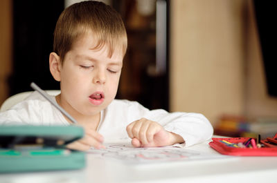 Cute and smiling cucasian boy draws with colored pencils while sitting at the table. 