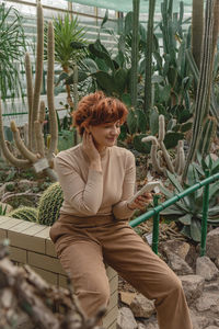 A beautiful plus size girl uses her smartphone sitting among the green plants of the greenhouse.