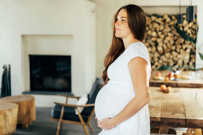 A pregnant woman in a white dress hugs her stomach with her hands.