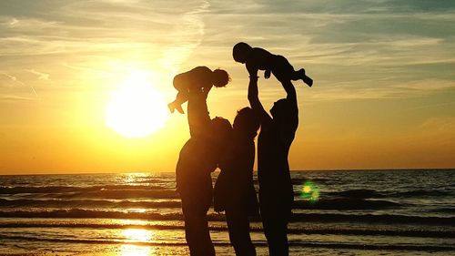 Silhouette women and man holding children and standing against sea during sunset