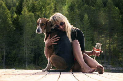 Young woman holding wineglass embracing dog on floorboard against trees