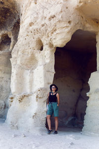 Woman traveler with a backpack and a hat stands next to a rock cave in the crimea in the summer