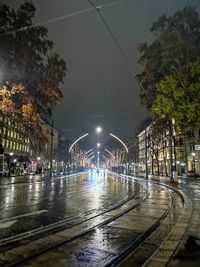 Illuminated railroad tracks by street against sky at night