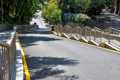 A new asphalt road descending down the slope of city street is flanked by metal railings along steps