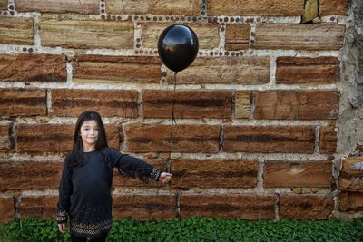 Portrait of smiling girl holding balloon while standing against brick wall