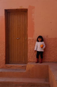 Full length portrait of cute girl holding pumpkin while standing by closed door