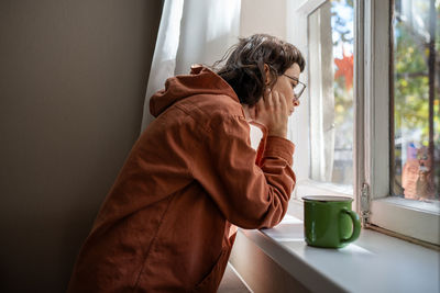 Introverted teen girl standing near window, looking at street indifferently, thinking of life